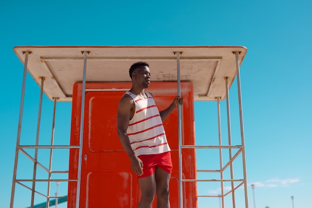 Jeune homme afro-américain regardant loin tout en se tenant sur la cabane de maître nageur à la plage contre le ciel bleu