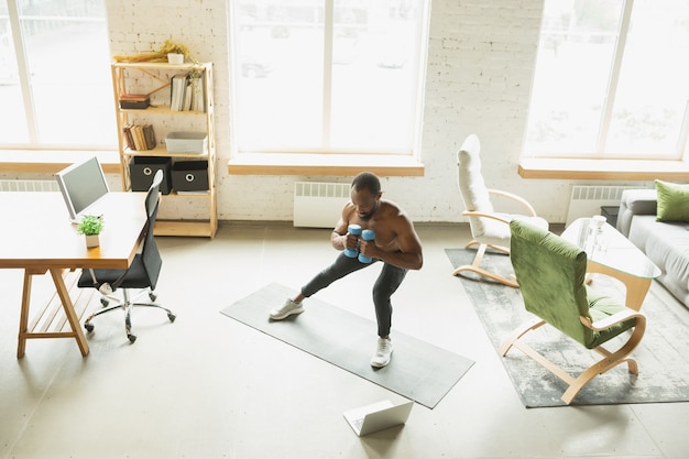 Jeune homme afro-américain s'entraînant à la maison pendant la quarantaine de l'épidémie de coronavirus, faisant des exercices de fitness, aérobie. Rester sportif pendant l'isolation. Bien-être, concept de mouvement. Asseyez-vous.