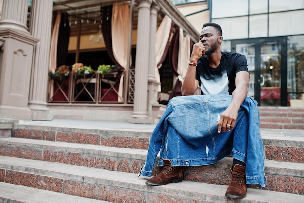 Jeune homme afro-américain sérieux et séduisant modèle moderne beau mec en veste de jeans assis sur la rue de la ville urbaine