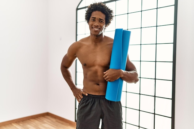 Jeune homme afro-américain souriant heureux tenant un tapis de yoga au gymnase