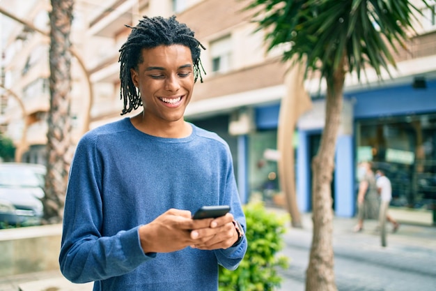 Jeune homme afro-américain souriant heureux en utilisant un smartphone dans la rue de la ville