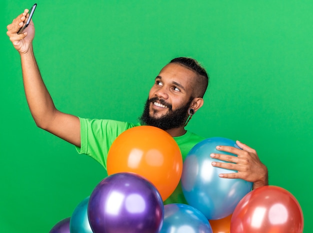 Un jeune homme afro-américain souriant portant un t-shirt vert debout derrière des ballons prend un selfie