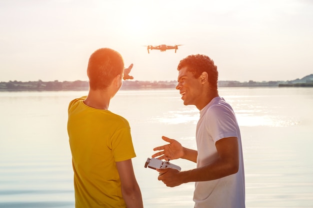 Jeune Homme Afro-américain Souriant Et Son Ami Caucasien Utilisant Un Drone Avec Contrôleur, Debout Près De La Rivière Au Coucher Du Soleil