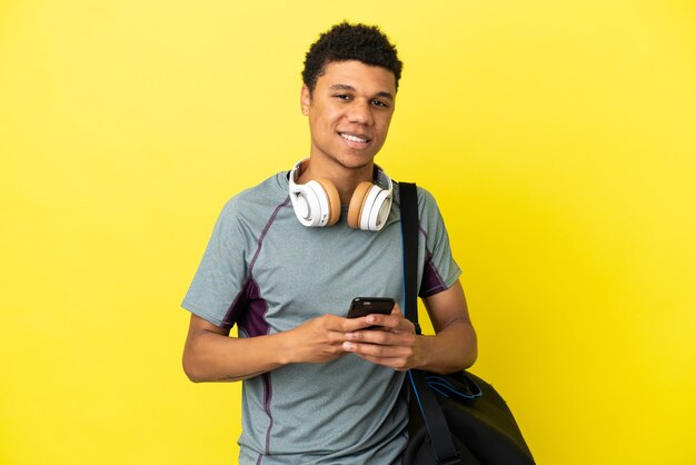 Jeune homme afro-américain de sport avec sac de sport isolé sur fond jaune envoyant un message avec le mobile