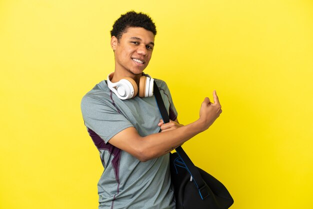 Jeune homme afro-américain de sport avec sac de sport isolé sur fond jaune pointant vers l'arrière