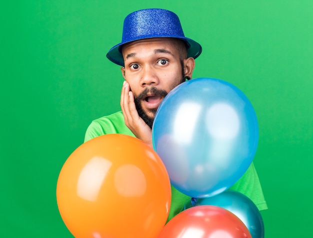 Jeune homme afro-américain surpris portant un chapeau de fête debout derrière des ballons mettant la main sur la joue isolée sur un mur vert