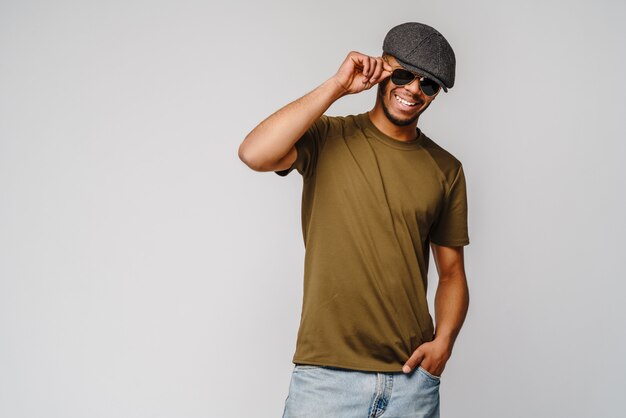 Jeune homme afro-américain en t-shirt vert portant des lunettes de soleil et une casquette sur un mur gris clair avec un espace vide