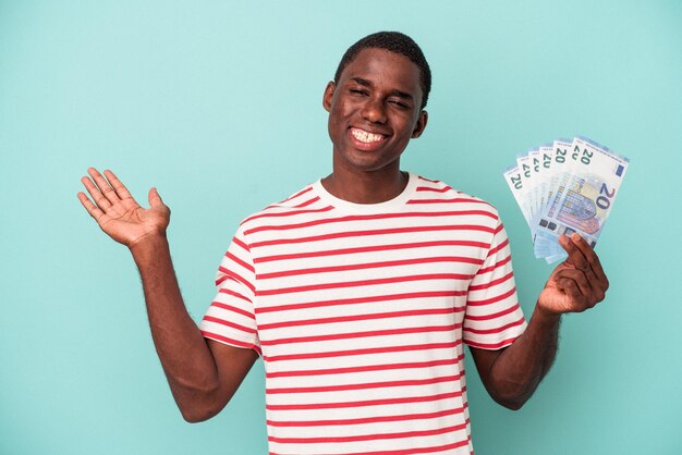 Jeune homme afro-américain tenant un billet de banque isolé sur fond bleu montrant un espace de copie sur une paume et tenant une autre main sur la taille