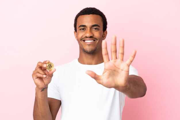 Jeune homme afro-américain tenant un Bitcoin sur fond rose isolé comptant cinq avec les doigts