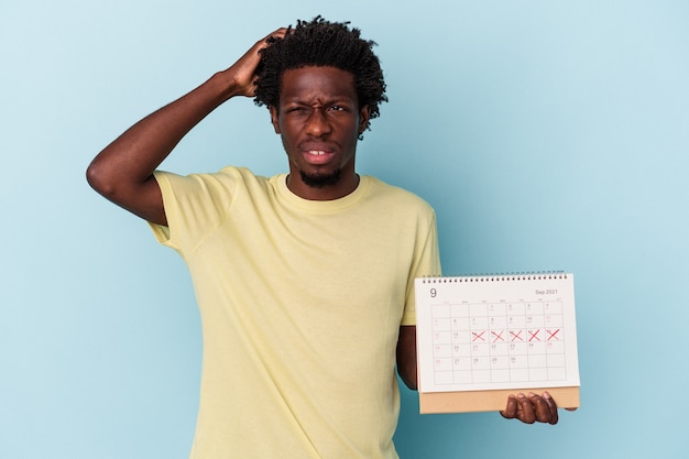 Jeune homme afro-américain tenant un calendrier isolé sur fond bleu étant choqué, elle s'est souvenue d'une réunion importante.