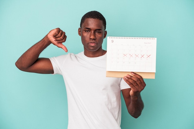 Jeune homme afro-américain tenant un calendrier isolé sur fond bleu montrant un geste d'aversion, les pouces vers le bas. Notion de désaccord.