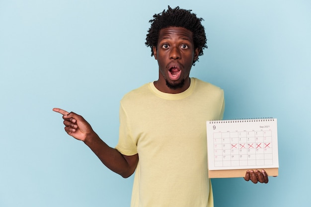 Jeune homme afro-américain tenant un calendrier isolé sur fond bleu pointant vers le côté