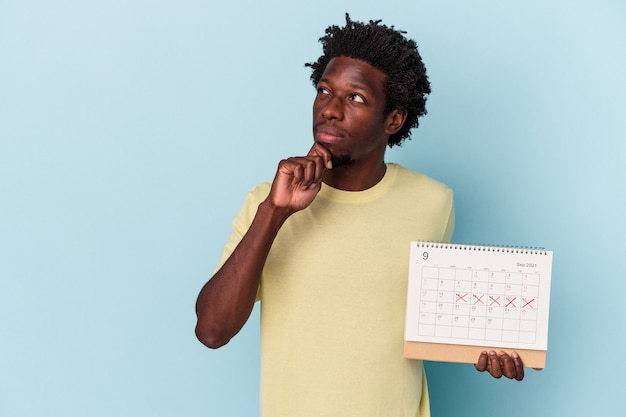 Jeune homme afro-américain tenant un calendrier isolé sur fond bleu regardant de côté avec une expression douteuse et sceptique.