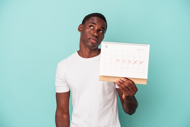 Jeune homme afro-américain tenant un calendrier isolé sur fond bleu rêvant d'atteindre des objectifs et des buts