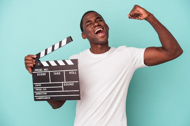 Jeune Homme Afro-américain Tenant Un Clap Isolé Sur Fond Bleu Levant Le Poing Après Une Victoire, Concept Gagnant.