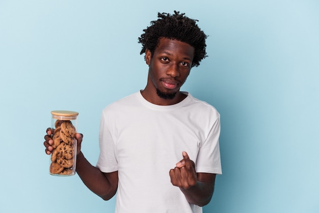 Jeune homme afro-américain tenant des cookies aux pépites de chocolat isolés sur fond bleu pointant du doigt vers vous comme s'il vous invitait à vous rapprocher.