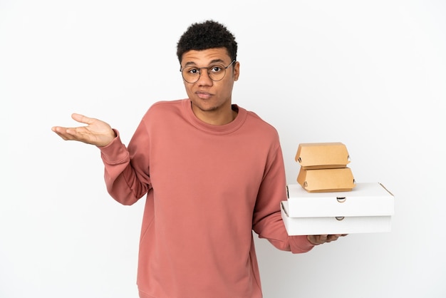 Jeune homme afro-américain tenant un hamburger et des pizzas isolés sur fond blanc ayant des doutes tout en levant les mains