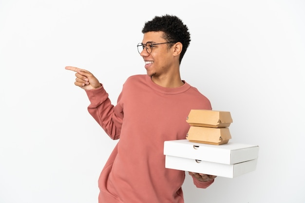 Jeune homme afro-américain tenant un hamburger et des pizzas isolés sur fond blanc, pointant le doigt sur le côté et présentant un produit