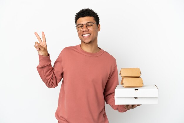 Jeune homme afro-américain tenant un hamburger et des pizzas isolés sur fond blanc souriant et montrant le signe de la victoire