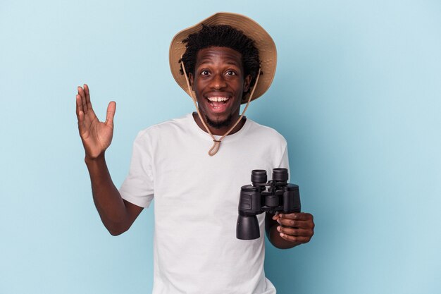 Jeune homme afro-américain tenant des jumelles isolées sur fond bleu recevant une agréable surprise, excité et levant les mains.