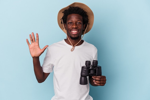 Jeune homme afro-américain tenant des jumelles isolées sur fond bleu souriant joyeux montrant le numéro cinq avec les doigts.