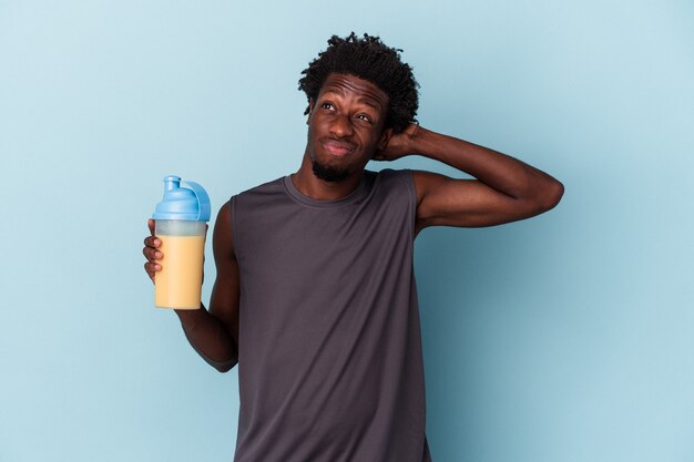 Jeune homme afro-américain tenant un milkshake protéiné isolé sur fond bleu touchant l'arrière de la tête, pensant et faisant un choix.