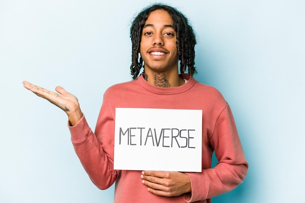 Jeune homme afro-américain tenant une pancarte métaverse isolée sur fond bleu montrant un espace de copie sur une paume et tenant une autre main sur la taille