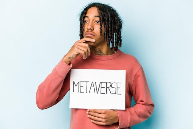 Jeune homme afro-américain tenant une pancarte métaverse isolée sur fond bleu regardant de côté avec une expression douteuse et sceptique