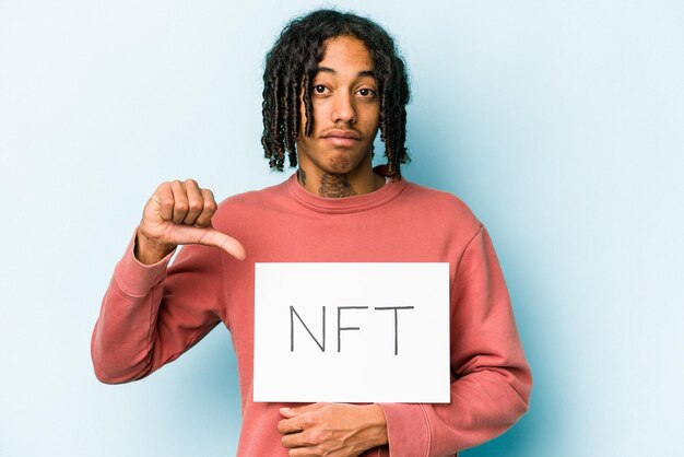 Photo jeune homme afro-américain tenant une pancarte nft isolée sur fond bleu montrant un geste d'aversion pouce vers le bas concept de désaccord