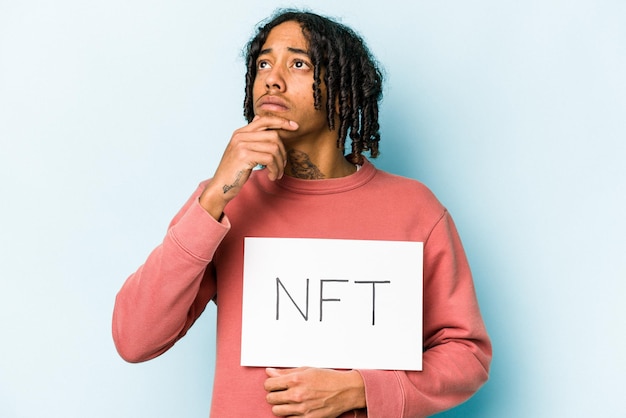 Jeune homme afro-américain tenant une pancarte NFT isolée sur fond bleu regardant de côté avec une expression douteuse et sceptique