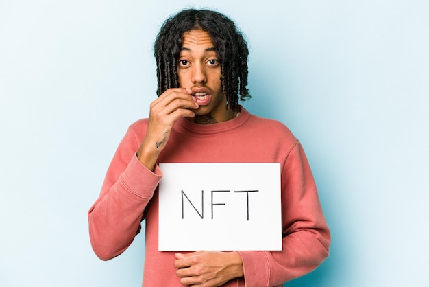 Photo jeune homme afro-américain tenant une pancarte nft isolée sur fond bleu se rongeant les ongles nerveux et très anxieux