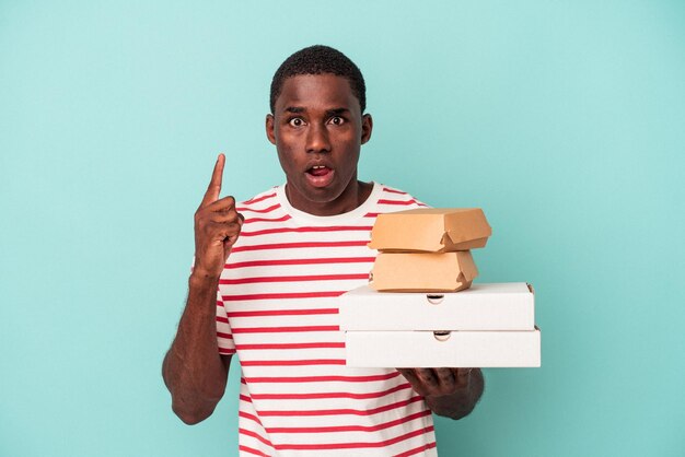 Jeune homme afro-américain tenant des pizzas et des hamburgers isolés sur fond bleu ayant une excellente idée de concept de créativité