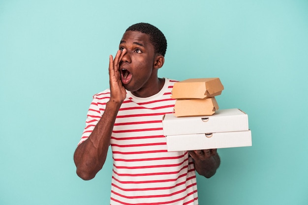 Jeune homme afro-américain tenant des pizzas et des hamburgers isolés sur fond bleu criant et tenant la paume près de la bouche ouverte.