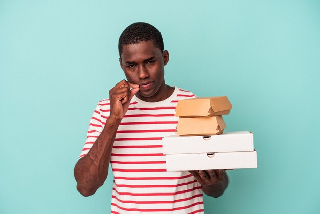 Jeune homme afro-américain tenant des pizzas et des hamburgers isolés sur fond bleu avec les doigts sur les lèvres gardant un secret.