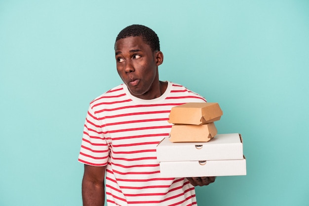 Jeune homme afro-américain tenant des pizzas et des hamburgers isolés sur fond bleu hausse les épaules et ouvre les yeux confus