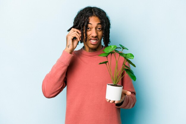 Jeune homme afro-américain tenant une plante isolée sur fond bleu couvrant les oreilles avec les mains