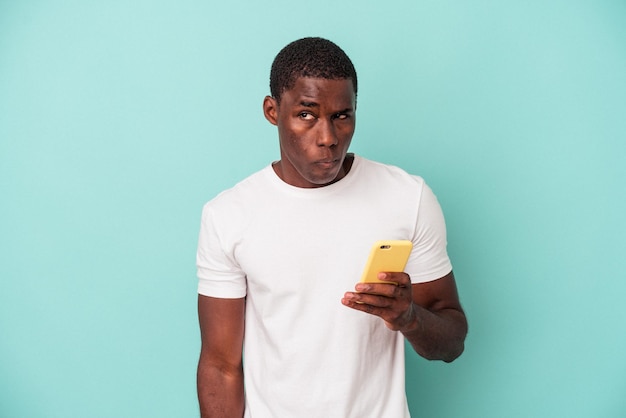 Jeune homme afro-américain tenant un téléphone portable isolé sur fond bleu confus se sent douteux et incertain