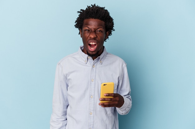 Jeune homme afro-américain tenant un téléphone portable isolé sur fond bleu criant très en colère et agressif.