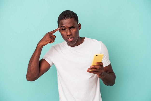 Jeune homme afro-américain tenant un téléphone portable isolé sur fond bleu montrant un geste de déception avec l'index.