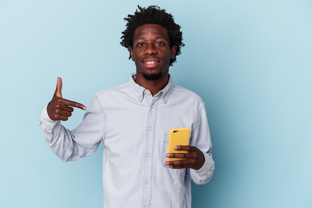 Jeune homme afro-américain tenant un téléphone portable isolé sur fond bleu personne pointant à la main vers un espace de copie de chemise, fier et confiant
