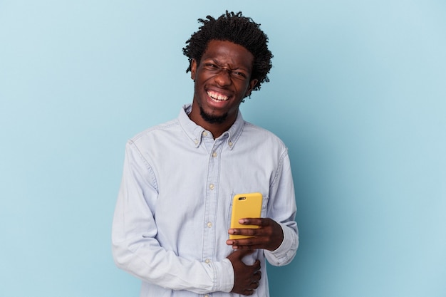 Jeune homme afro-américain tenant un téléphone portable isolé sur fond bleu en riant et en s'amusant.