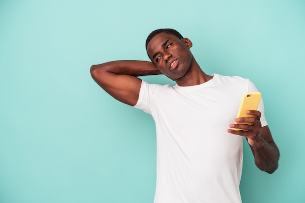 Jeune homme afro-américain tenant un téléphone portable isolé sur fond bleu touchant l'arrière de la tête, pensant et faisant un choix.