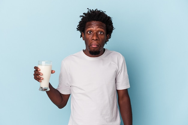 Jeune homme afro-américain tenant un verre de lait isolé sur fond bleu hausse les épaules et ouvre les yeux confus.