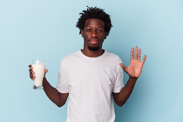 Jeune homme afro-américain tenant un verre de lait isolé sur fond bleu souriant joyeux montrant le numéro cinq avec les doigts.