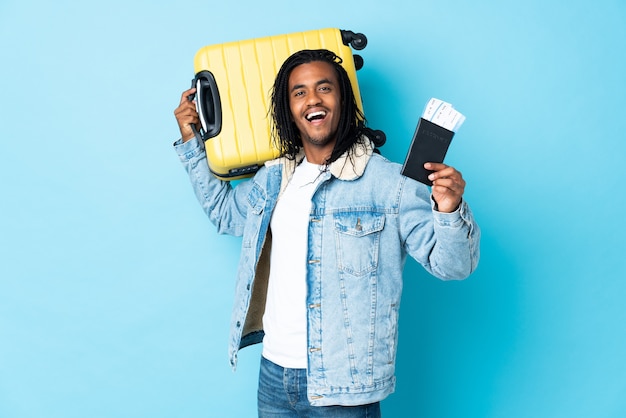 Jeune homme afro-américain avec des tresses isolé sur fond bleu en vacances avec valise et passeport