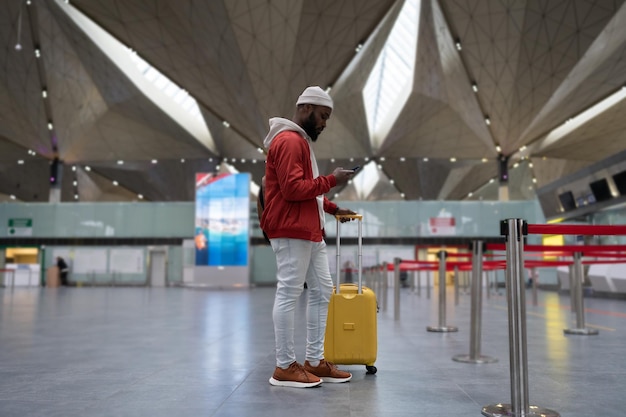 Jeune homme afro-américain utilisant un smartphone voyageant avec des bagages à l'intérieur du terminal de l'aéroport