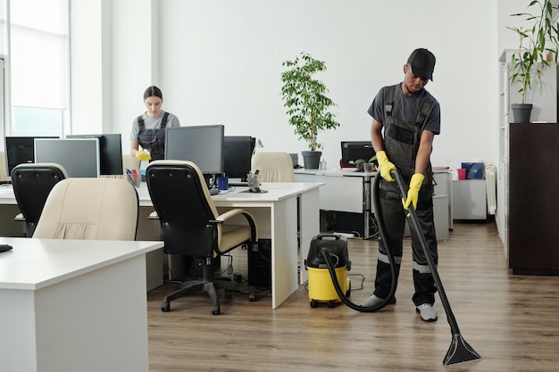 Jeune homme afro-américain en vêtements de travail nettoyant le sol d'un bureau contemporain