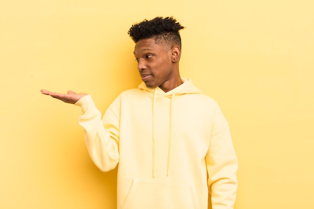 Jeune homme afro noir se sentant heureux et souriant avec désinvolture en regardant un objet ou un concept tenu sur la main sur le côté