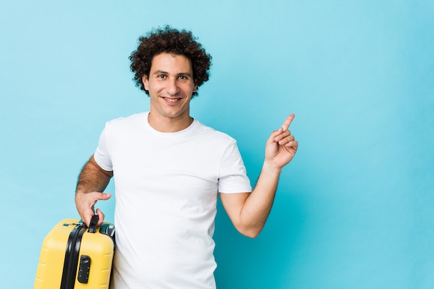 Jeune homme d'âge moyen tenant une valise souriant pointant joyeusement avec l'index loin.