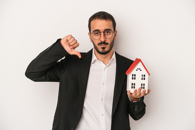 Jeune homme d'agent immobilier tenant une maison modèle isolée sur fond blanc montrant un geste d'aversion, les pouces vers le bas. Notion de désaccord.